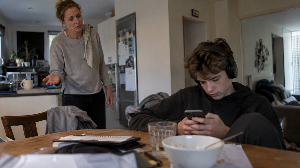 Tonåring tittar på mobilen vid köksbordet medan en vårdnadshavare försöker få kontakt och ge en instruktion.